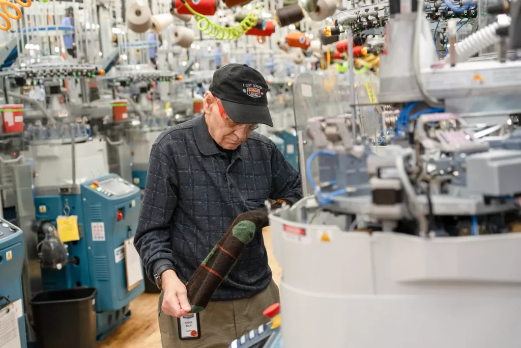 Operario con gorra y camisa negra de Darn Tough inspeccionando un calcetín marrón y verde.