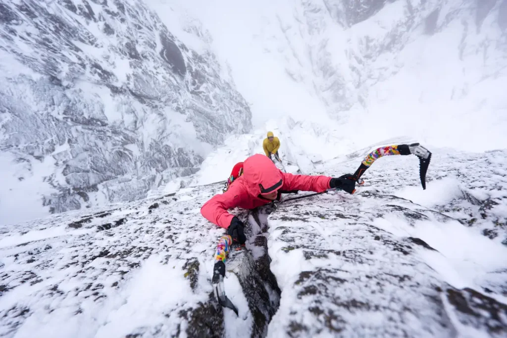 En una montaña nevada dos escaladores escalando en hielo el primero con dos piolets y chaqueta rosa y el que asegura con chaqueta amarilla