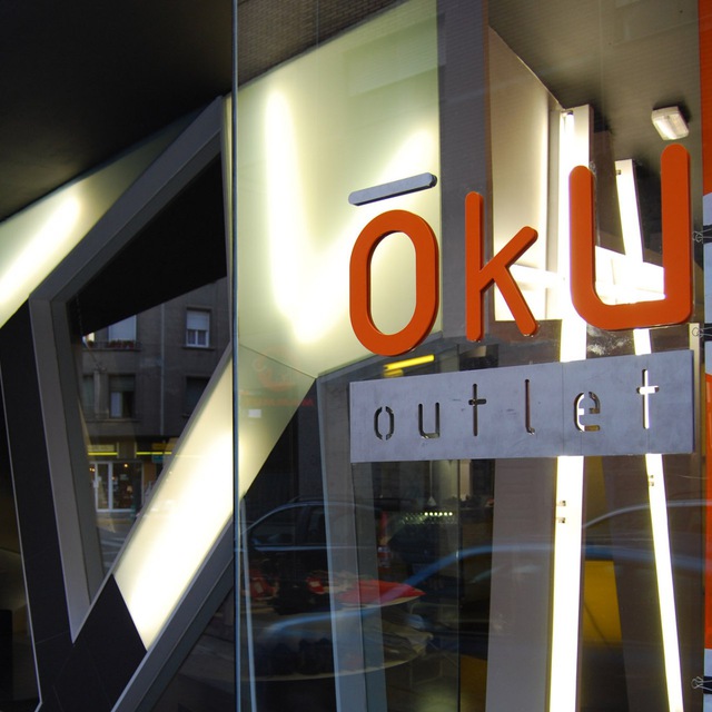 Cartel OkUoutlet Cartel de OkUoutlet con oku en letras naranja y outlet en negativo y metal. Al fondo se ven las luces de la fachada.