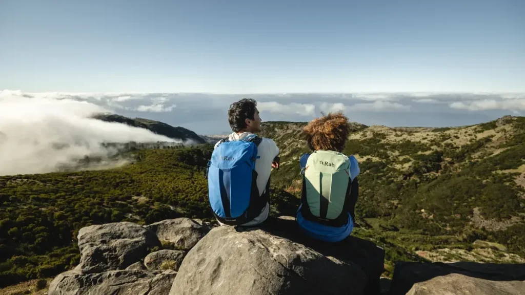 Un chico y una chica sentados en una roca en lo alto de una montaña con una mochila azul el chico y verde la chica con el cielo al fondo y montañas.