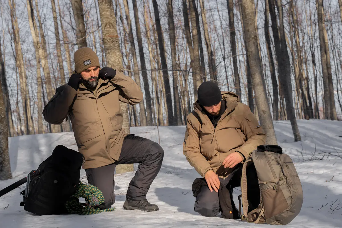 Dos hombres arrodillados sobre la nieve y unos arboles al fondo los dos llevan chaqueta marrón y pantalón negro con mochila