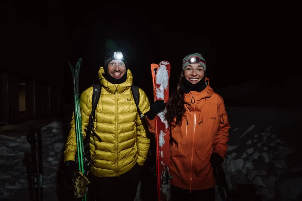 Dos esquiadores de noche con esquis en la mano derecha y frontales el chico lleva un plumas amarillo y la chica una chaqueta naranja 