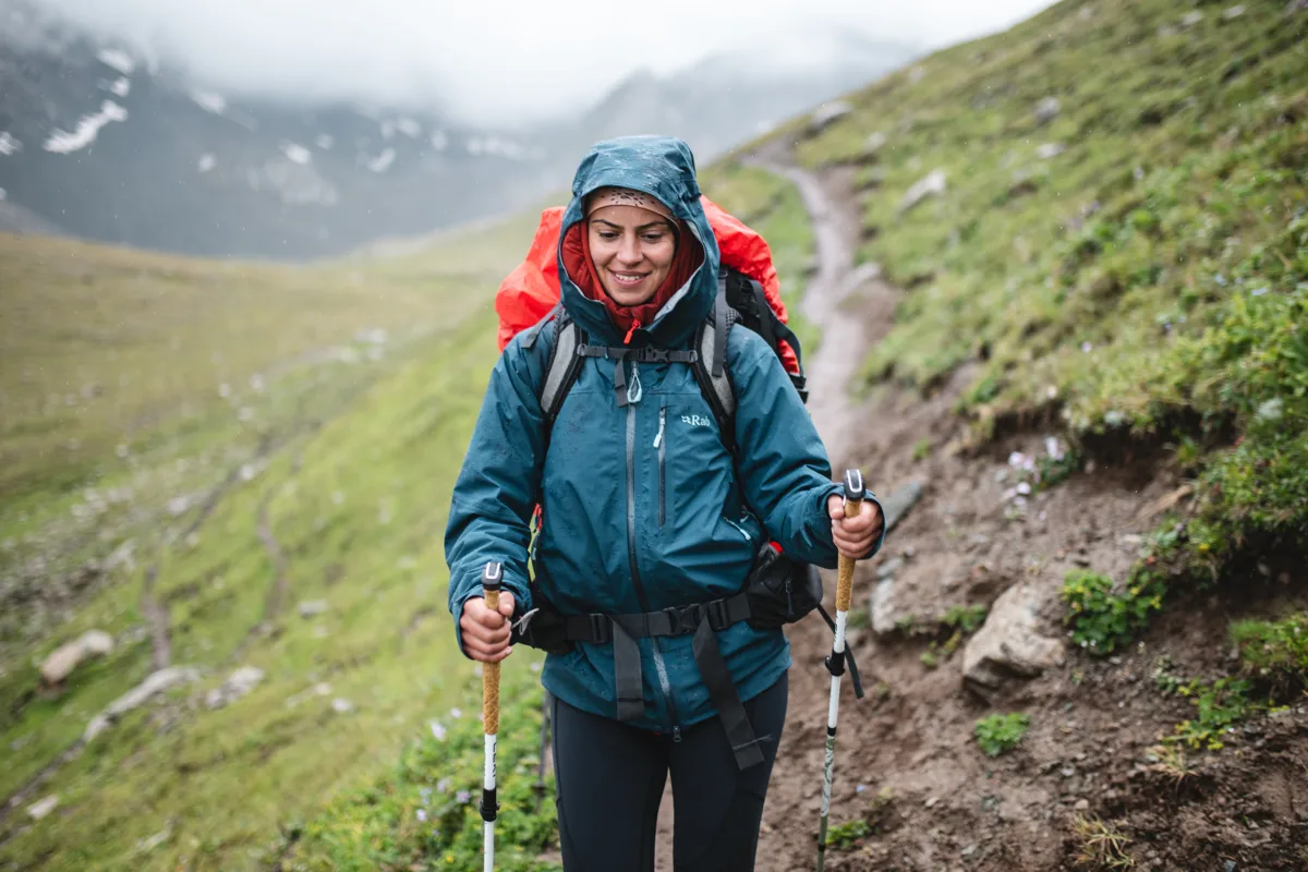 Senderista, caminando por un sendero en la montaña con chaqueta Rab azul bastones y mochila naranja.