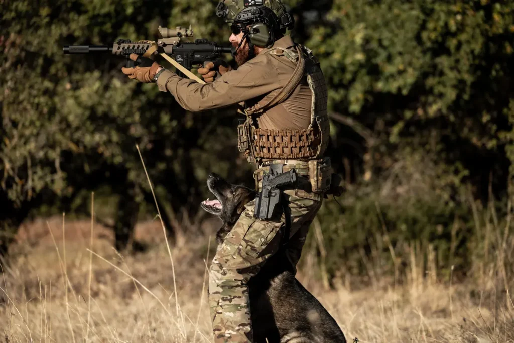 soldado con rifle en un bosque con pantalón de camuflaje y pistola negra con jersey marrón empuñando un rifle con casco y gafas.