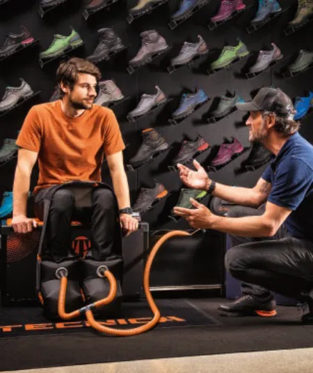 Fondo negro con zapatillas y dos chicos uno con camiseta naranja con un aparato negro en los pies y al lado un chico con gorra