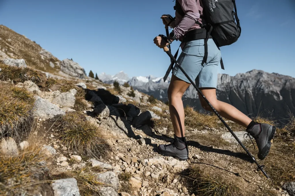 Senderista en pantalones cortos azules con bastones subiendo por la montaña con botas grises y e cielo de fondo.