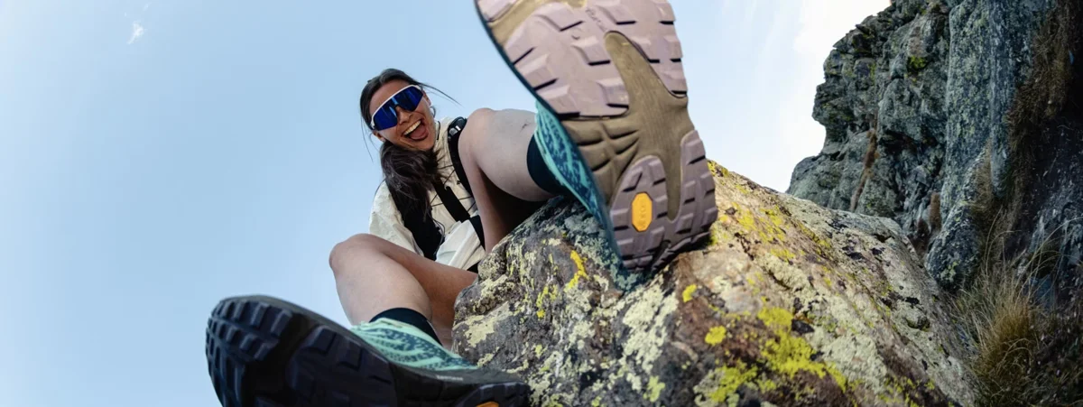 Chica sentada en una roca con gafas de sol negras. Vista desde abajo con zapatilla topo y suela vibram gris.