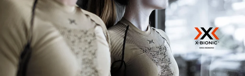 Un chico y una chica con ropa térmica marrón y la fondo unas montañas y el logo xbionic en negro y naranja.