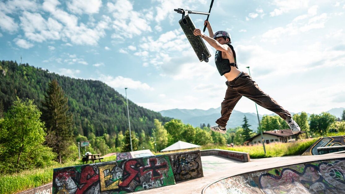 Teen executing a high freestyle scooter jump in Bergamo, Italy, with scenic mountain backdrop in daylight.