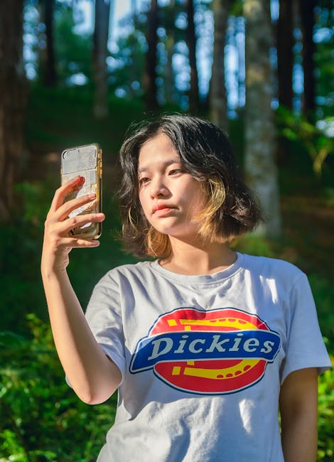 Moda Urbana A young woman taking a selfie with a smartphone in a sunlit forest, wearing a casual Dickies t-shirt.