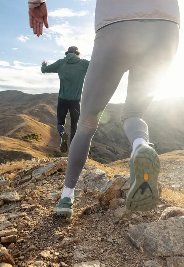 Un chico con pantalón negro y chaqueta verde y una chica corriendo con mallas gris, calcetines blancos y zapatillas verdes. 